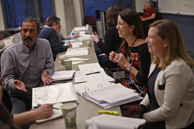 A group of four people sitting at a table engaged in a discussion, with papers and notebooks spread out before them. Two women are smiling, and a man is speaking. Other meeting participants are in the background.